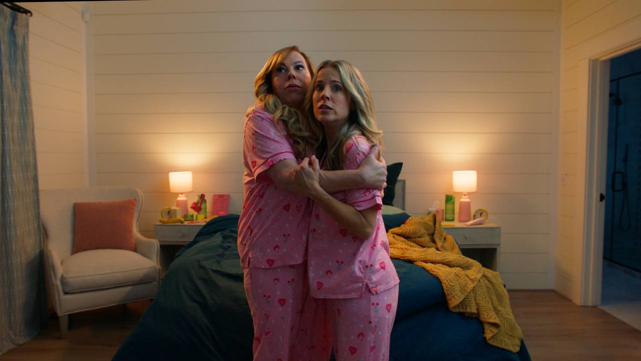Two women in matching pink pajamas embrace in a warmly lit bedroom.