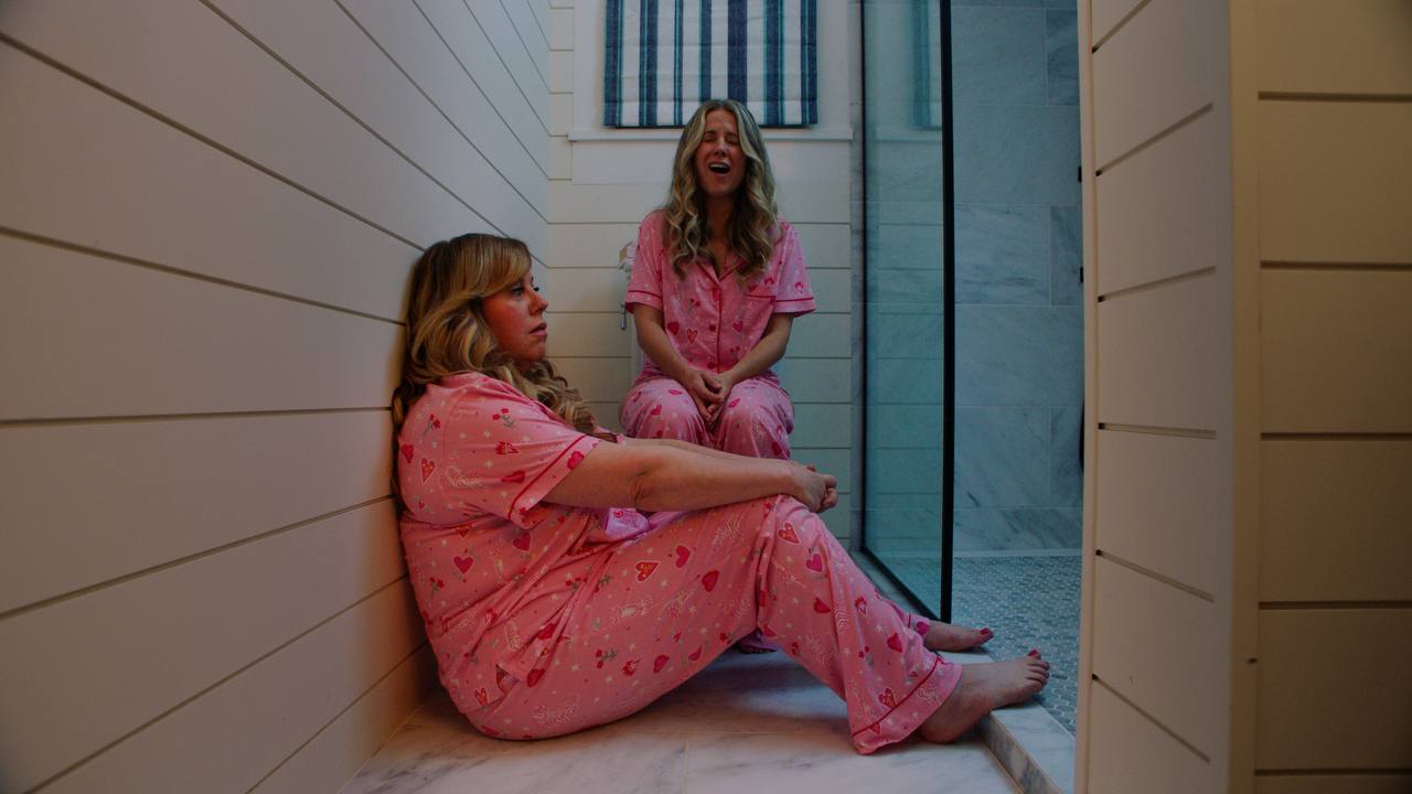 Two women in pink pajamas sit in a narrow bathroom hallway.